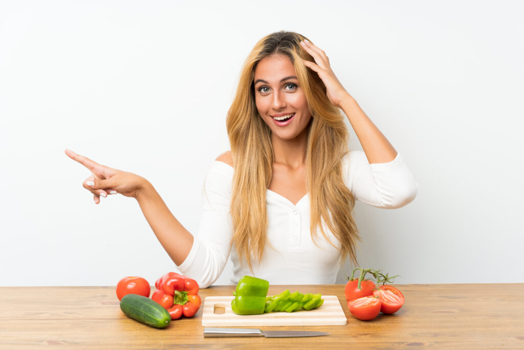 young blonde woman with vegetables table surprised pointing finger side